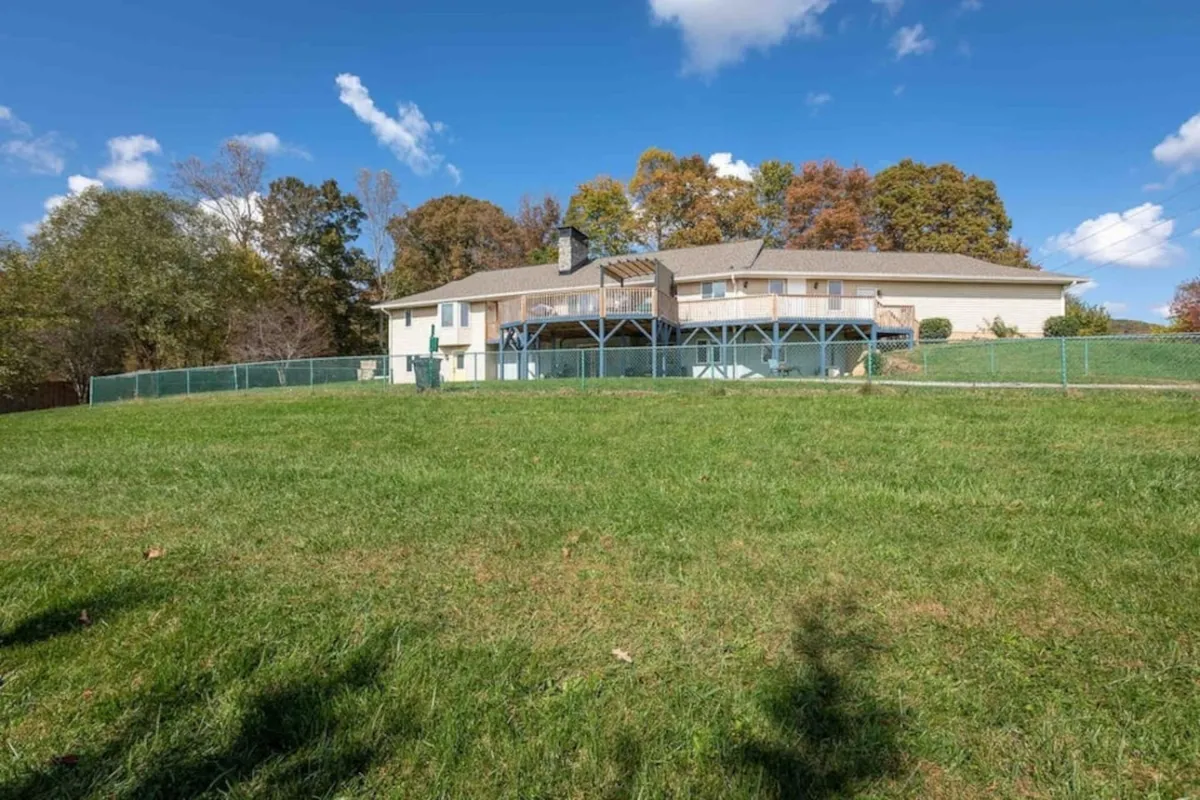 A welcoming view of the house's exterior, featuring a spacious deck overlooking an expansive lawn. The scene is inviting, with the sun setting beyond the deck, and hints of family gatherings, barbecues, and children playing outdoors. The backdrop hints at the surrounding mountain landscape, emphasizing a sense of tranquility and retreat.