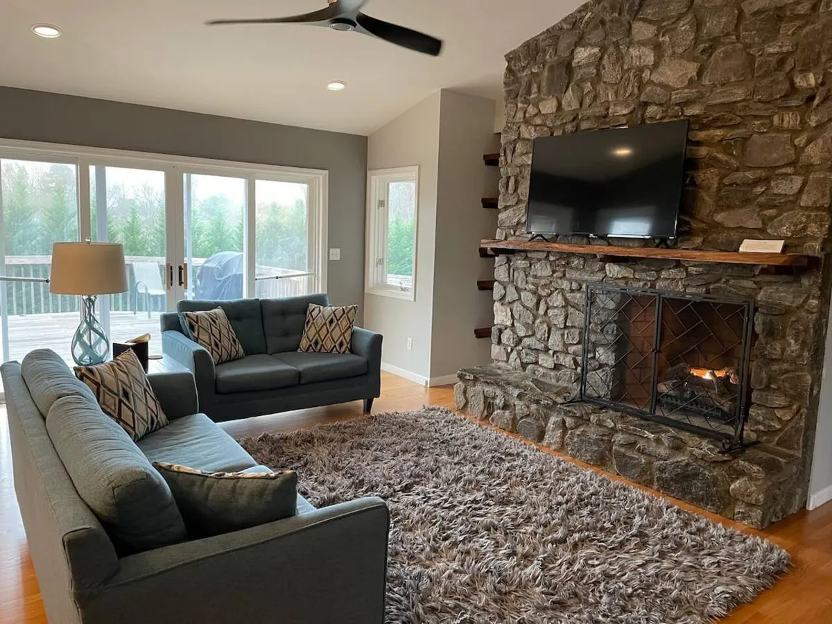 A spacious, inviting living room with a large stone fireplace as the focal point. Plush sofas are arranged to encourage conversation and comfort, while large windows let in abundant natural light and offer mountain views. The overall atmosphere is cozy and perfect for gatherings, with plenty of seating for family and friends.