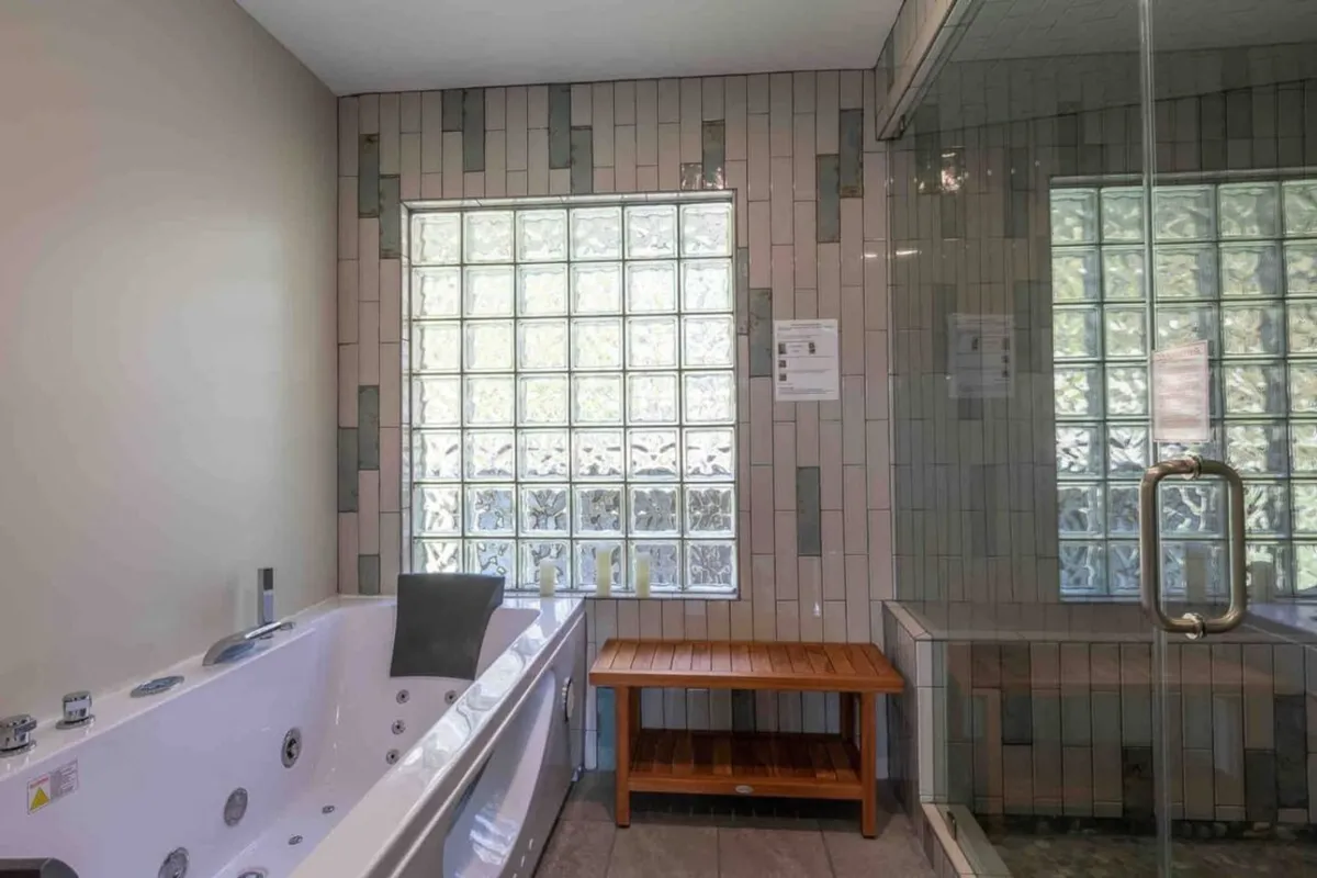 A serene spa-like bathroom featuring a two-person Jacuzzi tub set beneath a glass block window, with a wooden bench nearby adding warmth and comfort to the space. Natural light streams in, highlighting the tranquil atmosphere perfect for relaxation.