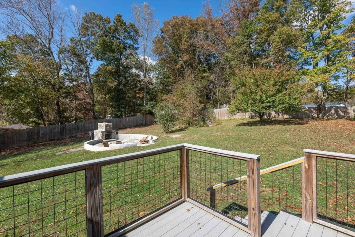 A spacious backyard with a fire pit surrounded by chairs, bordered by lush green lawn and mature trees, inviting for outdoor gatherings and play. The large open area is perfect for both energetic kids and relaxing adults, with natural scenery enhancing the sense of retreat.