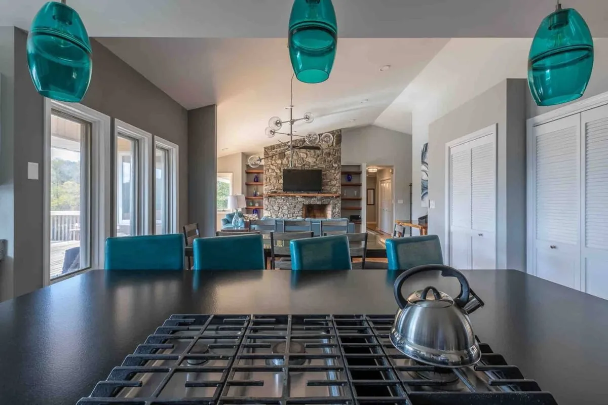 A stylish, modern kitchen highlighted by teal pendant lights, a large kitchen island with bar seating, and a stone fireplace in the background. The space is open and inviting, with ample countertops and room for multiple people to cook or gather. The decor blends warmth and contemporary touches, creating a vibrant atmosphere for culinary activities.
