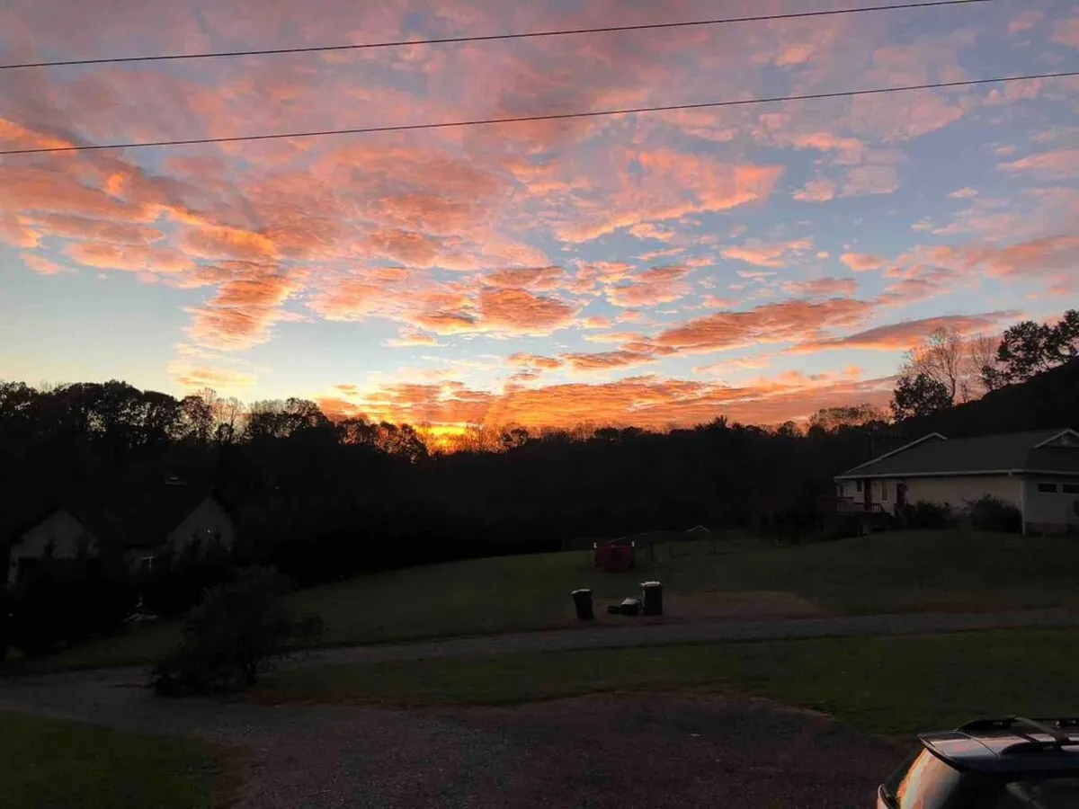 A breathtaking view of the sunset sky over silhouetted trees and rolling hills, with the horizon stretching in the distance. The tranquil setting evokes a serene mountain atmosphere, suggesting the beauty of the area surrounding the property.