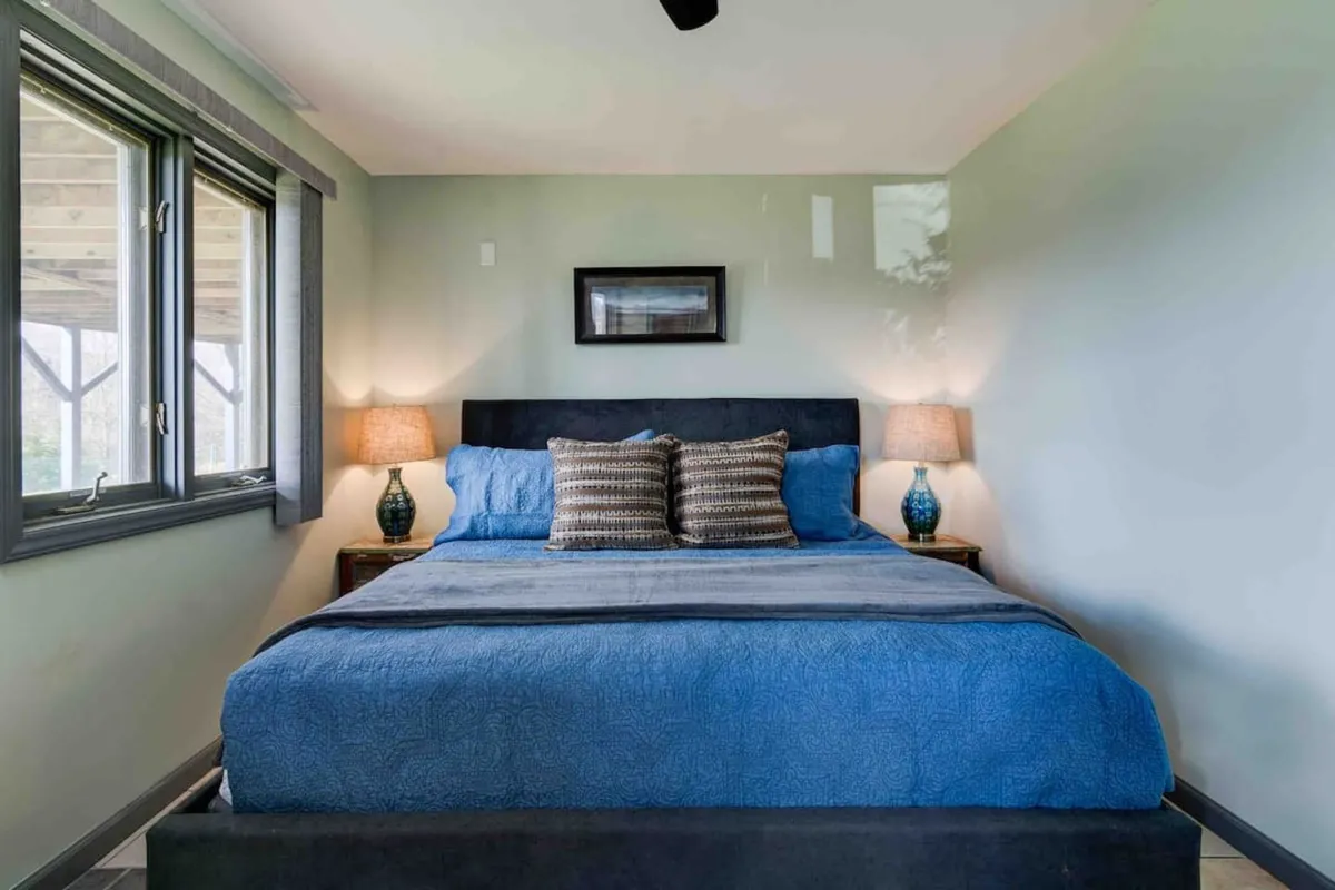 A cozy bedroom featuring a bed with blue bedding, decorative pillows, and table lamps on either side. Large windows invite natural light, creating a restful and serene environment ideal for reading or unwinding.