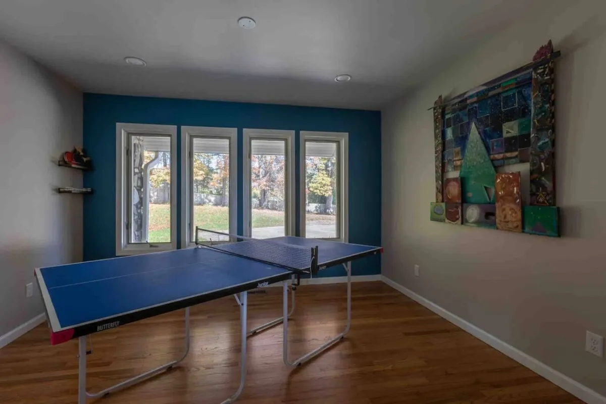 A bright, energetic game space with a ping pong table as the centerpiece, large windows providing views and natural light, and colorful artwork adding a playful touch. The room is designed for dynamic social gatherings and active fun.