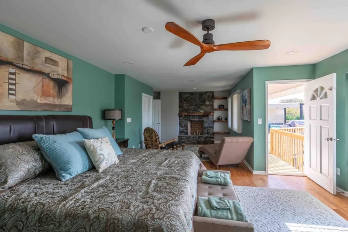 A sophisticated bedroom with a bed adorned with decorative pillows, tasteful artwork, a cozy fireplace, and an open door leading to the outdoors. The ambiance is serene, inviting guests to unwind by the fire or enjoy mountain views with morning coffee.