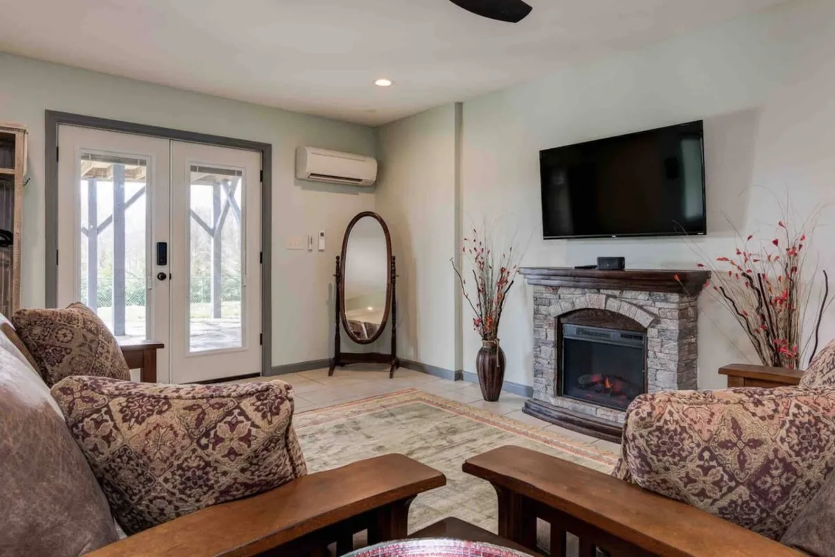 A stylish living area featuring a cozy fireplace, comfortable seating, a TV, and decorative plants. The space exudes warmth and is designed for socializing or relaxing, with large windows likely offering views of the surrounding landscape.