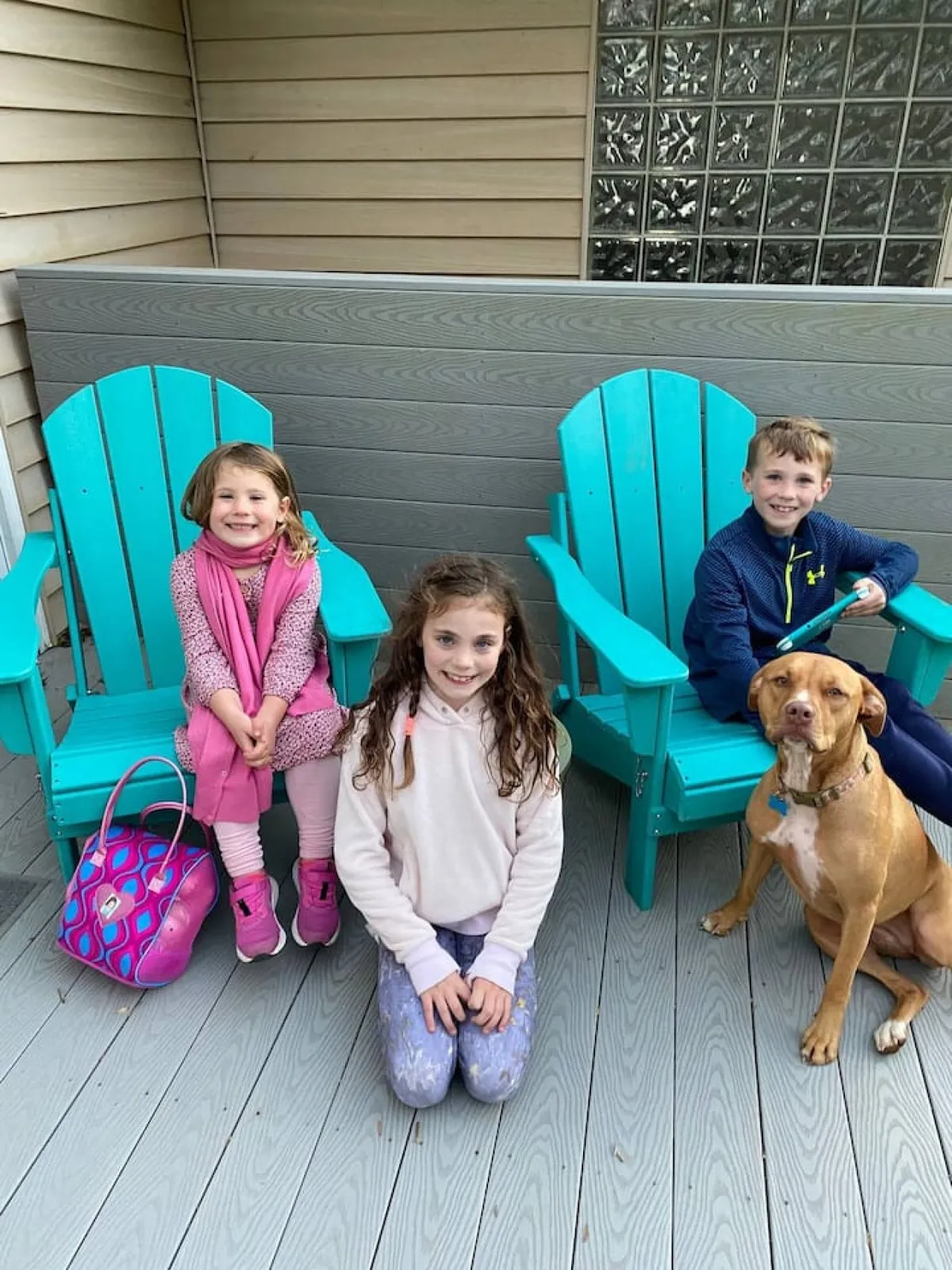 Children are seated in colorful chairs outdoors alongside a dog, enjoying a relaxed moment in the sunshine. The setting is cheerful and playful, hinting at safe, enjoyable outdoor spaces for kids and pets to explore and unwind.
